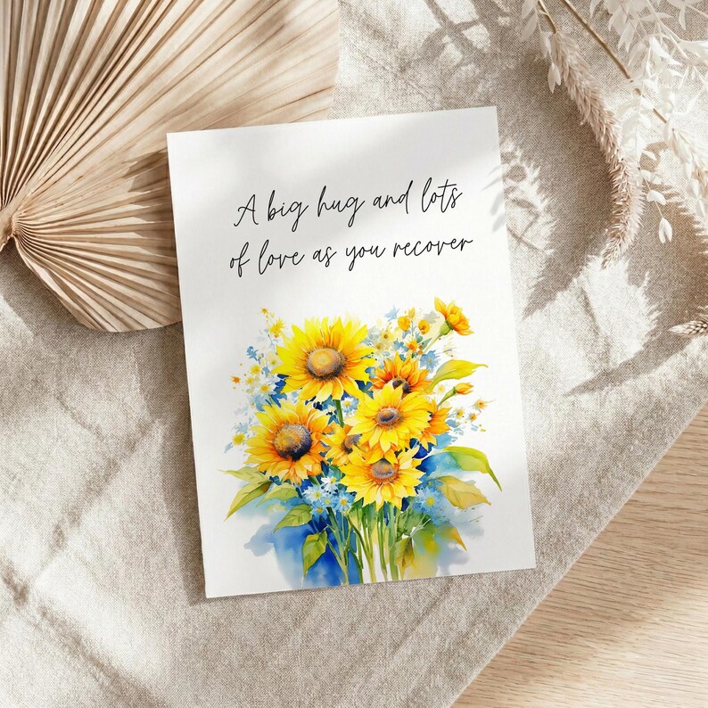 A greeting card with a floral design and a message about love, placed on a beige cloth next to a fan and some dried flowers.