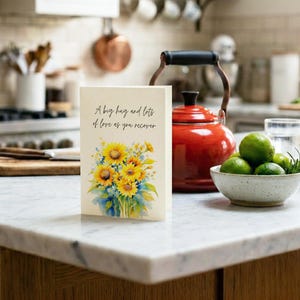 A card with a floral design and a message about love is displayed on a kitchen counter, accompanied by a red kettle, a bowl of fruit, and a cutting board.