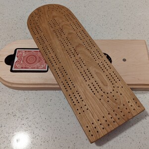 May include: A wooden cribbage board with a light-colored base and a darker, rounded top piece featuring rows of drilled holes. A deck of playing cards with a red patterned back is tucked into a recessed compartment within the base of the board.