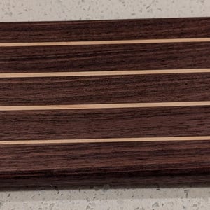 May include: A rectangular wooden serving board with a handle, featuring dark brown and light wood stripes. A white, oval-shaped bowl sits on the board. This item is suitable for serving appetizers or small dishes.