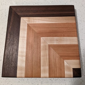 Könnte beinhalten: Ein quadratisches Schneidebrett aus Holz mit einem dunkelbraunen Rand und einem hellbraunen Chevron-Muster in der Mitte.