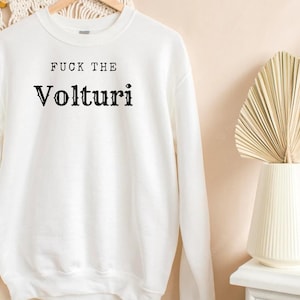 May include: White sweatshirt with the text "FUCK THE Volturi" in black lettering. The sweatshirt is hanging on a wooden hanger. A decorative vase with dried palm leaves is in the background.