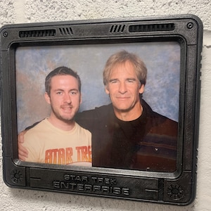 May include: A framed photograph featuring two men, set within a black, retro-style frame. The frame is labeled "STAR TREK ENTERPRISE" at the bottom. One man wears a shirt with "STAR TREK" printed on it.