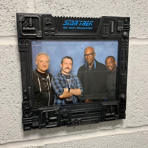 Puede incluir: Marco de fotos negro de Star Trek: The Next Generation con una foto de cuatro hombres. El marco tiene un diseño geométrico detallado. El logotipo de Star Trek es azul. El marco está montado en una pared de ladrillos blancos.
