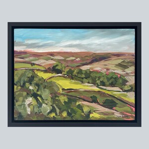 May include: An original landscape painting in a black frame. The artwork depicts rolling hills in shades of green, brown, and pink under a blue sky with white clouds. The artist's signature is visible in the lower right corner.