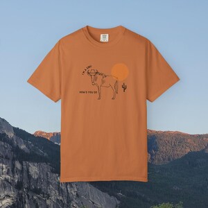 Puede incluir: Una camiseta de manga corta color naranja quemado con una ilustración de líneas negras de un ñu con sombrero de vaquero. El texto dice I'M A GNU y HOW'D YOU DO, junto a un círculo naranja sólido que representa el sol y un pequeño cactus.