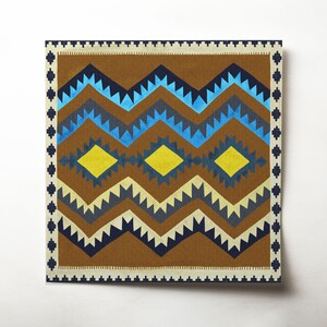 Op de afbeelding: Vierkante textiel met een geometrisch patroon in bruin, blauw, geel en crème. Het ontwerp heeft zigzaglijnen en ruitvormen, omlijst door een rand met kleine, zich herhalende elementen.