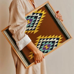 Könnte beinhalten: Ein gerahmter Druck mit einem geometrischen Muster in Braun-, Gelb-, Blau- und Violetttönen. Das Muster erinnert an einen traditionellen Teppich oder Stoff.