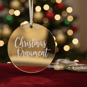 Puede incluir: Adorno navideño dorado con las palabras "Christmas Ornament" en escritura blanca. El adorno es redondo y tiene una cinta blanca para colgarlo. El fondo es un árbol de Navidad borroso con luces rojas y doradas.