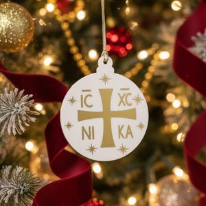 以下が含まれることがあります： 金色の十字架とギリシャ語のテキストが入った白いクリスマスオーナメント。オーナメントは丸く、星のアクセントがあり、金の紐から吊り下げられています。背景には、赤いリボンと金と赤のオーナメントが付いたクリスマスツリーがあります。