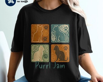May include: Black t-shirt featuring a four-panel design with cat silhouettes, yarn, and paw prints in orange, teal, cream, and brown. The text "Purrl Jam" is printed below the panels in teal.