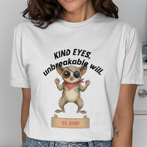 Puede incluir: Camiseta blanca con un tarsio de dibujos animados con un parche en el ojo y un pañuelo rojo. El texto "KIND EYES, unbreakable will." está encima del tarsio, y "BE KIND" está debajo. La camiseta es de material suave.