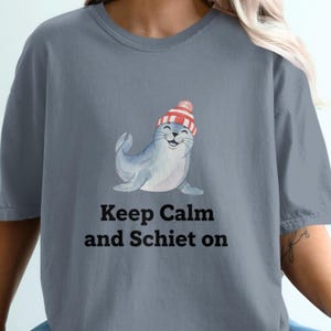 Könnte beinhalten: Blau-graues T-Shirt mit einem lächelnden Seehund, der eine rot-weiß gestreifte Mütze trägt. Der Text "Keep Calm and Schiet on" ist darunter gedruckt. Das Shirt ist ein lässiger, bequemer Stil.