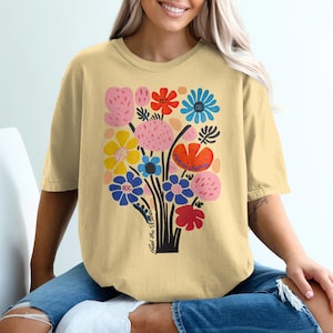 Op de afbeelding: Een lichtgeel T-shirt met een kleurrijk bloemenontwerp en de tekst "Find the Flower" gedrukt op de voorkant.