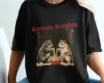 Könnte beinhalten: Schwarzes T-Shirt mit einem Grafikdruck von zwei Katzen, die Ramen-Nudeln essen. Der Text "Ramen Rumble" ist in Rot über den Katzen gedruckt.