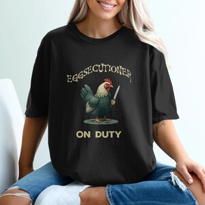 Può includere: Una maglietta nera con una grafica di un pollo che tiene un coltello. Il testo sulla maglietta recita "Eggsecutioner on Duty".