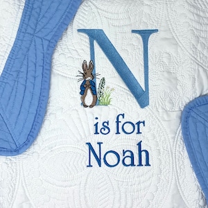 May include: A white baby blanket with a blue embroidered letter N, a Peter Rabbit illustration, and the text "is for Noah".