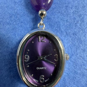 May include: An oval-shaped silver-tone quartz watch with a deep purple sunray dial. The watch face features silver Arabic numerals at the 12, 3, 6, and 9 o'clock positions, with silver hands and the text QUARTZ and JAPAN MOVT printed on the dial.