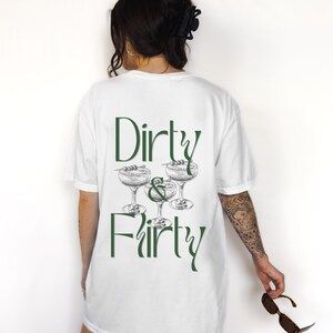 May include: White t-shirt with the text "Dirty & Thirty" in green lettering and a graphic of three martini glasses with olives.