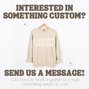 May include: A beige sweatshirt with the text "CUSTOM DESIGN" printed on the front. The text is in a white font. The sweatshirt is hanging on a wooden hanger against a white background. The text "INTERESTED IN SOMETHING CUSTOM?" is at the top of the image and the text "SEND US A MESSAGE! We'd love to work together to create something unique to you!" is at the bottom of the image.