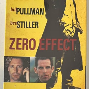 May include: A yellow and red VHS tape cover with the title "ZERO EFFECT" in red letters. The cover features a black and white image of a man in a coat, with the actors Bill Pullman and Ben Stiller listed below. The tagline reads "The World's Most Private Detective".