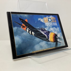 May include: A book titled "Ghosts Postcard Book" featuring a colourful World War II era fighter plane in flight against a blue sky with clouds. The book cover includes the text "20 Historic WWII Aircraft".