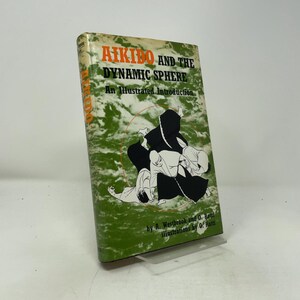 May include: A hardcover book titled Aikido and the Dynamic Sphere: An Illustrated Introduction by A. Westbrook and O. Ratti. The cover features a green and white marbled background with black ink illustrations of figures practicing martial arts.