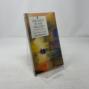 以下が含まれることがあります： ケネス・グレアム著、アラン・ベネット脚色の「The Wind in the Willows」というタイトルのハードカバー本。表紙には、水上のボートの水彩画イラストが描かれています。本は透明なアクリルスタンドに立てかけられています。