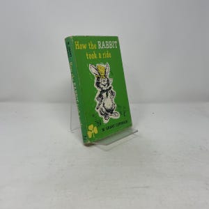 May include: A vintage children's book titled "How the RABBIT took a ride" with a green cover. The cover features a cartoon rabbit wearing a hat and holding a cane. The book is propped up on a clear stand.
