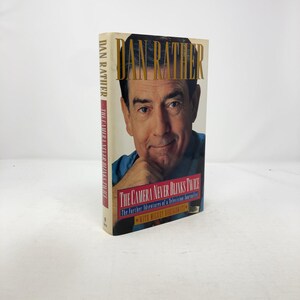 Pode incluir: Um livro de capa dura intitulado "The Camera Never Blinks Twice" de Dan Rather. A capa apresenta um retrato de Dan Rather com letras douradas. A lombada mostra o nome do autor e o título do livro.