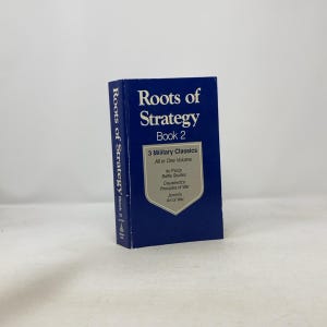 Op de afbeelding: Een marineblauw boek met de titel "Roots of Strategy Book 2". Het boek bevat de tekst "3 Military Classics" en vermeldt werken van du Picq, Clausewitz en Jomin. De rug toont ook de titel en het boeknummer.