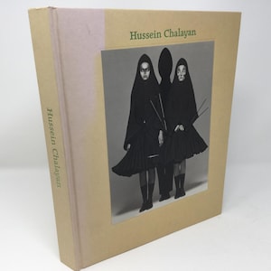 Pode incluir: Um livro intitulado "Hussein Chalayan" com uma fotografia em preto e branco de três pessoas vestidas de preto na capa.
