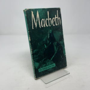 Puede incluir: Un libro antiguo titulado "Macbeth" con una cubierta verde y letras blancas. La portada presenta una imagen oscura de figuras y el texto "The Making of the Film". El libro se exhibe sobre un soporte transparente.