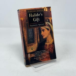 Op de afbeelding: Een hardcover boek met de titel "Halide's Gift" van Frances Kazan. De omslag toont een portret van een vrouw in traditionele kledij, tegen een achtergrond van sierlijke architecturale details. Het boek is een internationale bestseller.