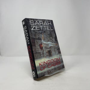 May include: A hardback book titled "Kingdom of Cages" by Sarah Zettel. The cover shows a woman suspended in the air, with a cityscape backdrop. The author's name is displayed at the top.
