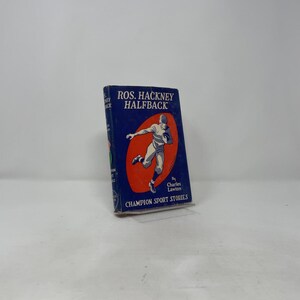 Puede incluir: Un libro antiguo titulado "Ros. Hackney Halfback" con una cubierta azul e ilustración de un jugador de fútbol americano. El libro es parte de la serie "Champion Sport Stories" y está escrito por Charles Lawton.