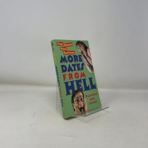 May include: A paperback book titled "More Dates From Hell" by Katherine Ann Samon. The cover is green with the title in large blue letters and images of surprised faces. The book is propped up on a clear plastic stand.