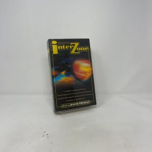 May include: A paperback book titled "The Best of Interzone" with a black cover and yellow lettering. The cover features a colorful space-themed illustration with planets and stars. Edited by David Pringle.