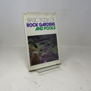 Könnte beinhalten: Ein Buch mit dem Titel "BASIC BOOK OF ROCK GARDENS AND POOLS" mit einem Foto eines Steingartens auf dem Einband. Das Buch ist weiß mit lila und grüner Schrift. Das Buch steht auf einem transparenten Kunststoffständer.