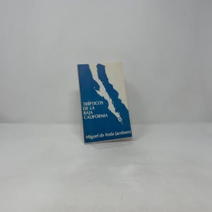 May include: A paperback book titled "Trípticos de la Baja California" by Miguel de Anda Jacobsen. The cover features a blue and white graphic of a coastline. The book is set against a white background.
