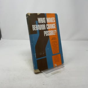 Puede incluir: Un libro de tapa dura titulado "WHAT MAKES BEHAVIOR CHANGE POSSIBLE?" con un diseño de portada marrón y azul. El libro está editado por Arthur Burton y presenta una lista de colaboradores. El libro se muestra en un soporte de plástico transparente.