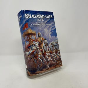 Peut inclure: Un livre à couverture rigide intitulé Bhagavad-Gita As It Is par A. C. Bhaktivedanta Swami Prabhupada. La couverture présente un ciel bleu avec une illustration détaillée de deux personnages sur un char doré tiré par quatre chevaux blancs.