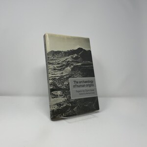 May include: A hardcover book titled "The archaeology of human origins" by Glynn Isaac, edited by Barbara Isaac. The cover features a black and white landscape image of mountains. The book is wrapped in clear plastic.