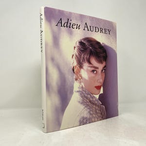 May include: A book titled "Adieu Audrey" with a photo of Audrey Hepburn on the cover. The photo shows her looking to the right, with a soft, pink background.