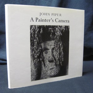 Puede incluir: Una cubierta de libro blanca con el título "A Painter's Camera" de John Piper. La cubierta presenta una fotografía en blanco y negro de una escultura de piedra de un rostro.