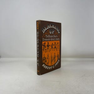 Puede incluir: Un libro de tapa dura marrón titulado "Blacking Up: The Minstrel Show in Nineteenth-Century America" de Robert C. Toll. La portada presenta texto e ilustraciones en naranja y blanco. El título está en letras grandes.