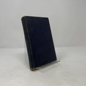 May include: A hardcover book with a solid dark navy blue cover and a black spine. The book is positioned upright on a clear acrylic stand, displaying its front cover and side spine. The surface of the cover appears matte and shows signs of wear.