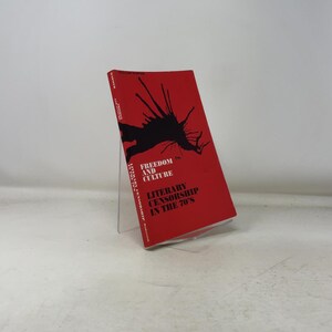 Puede incluir: Un libro rojo titulado "Freedom and Culture" con el subtítulo "Literary Censorship in the 70's". La portada presenta un diseño de mancha de tinta negra. El libro se exhibe en un soporte acrílico transparente.