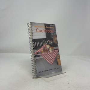 Può includere: Un libro di cucina rilegato a spirale intitolato "Midwest Old Threshers Cookbook" con un'immagine di cibo su una tovaglia a scacchi rossi e bianchi. Il libro di cucina è appoggiato su un supporto di plastica trasparente.