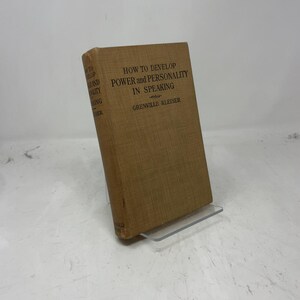 Può includere: Un libro vintage con copertina marrone, intitolato "How to Develop Power and Personality in Speaking" di Grenville Kleiser. Il libro è esposto su un supporto in acrilico trasparente, con testo nero sulla copertina.